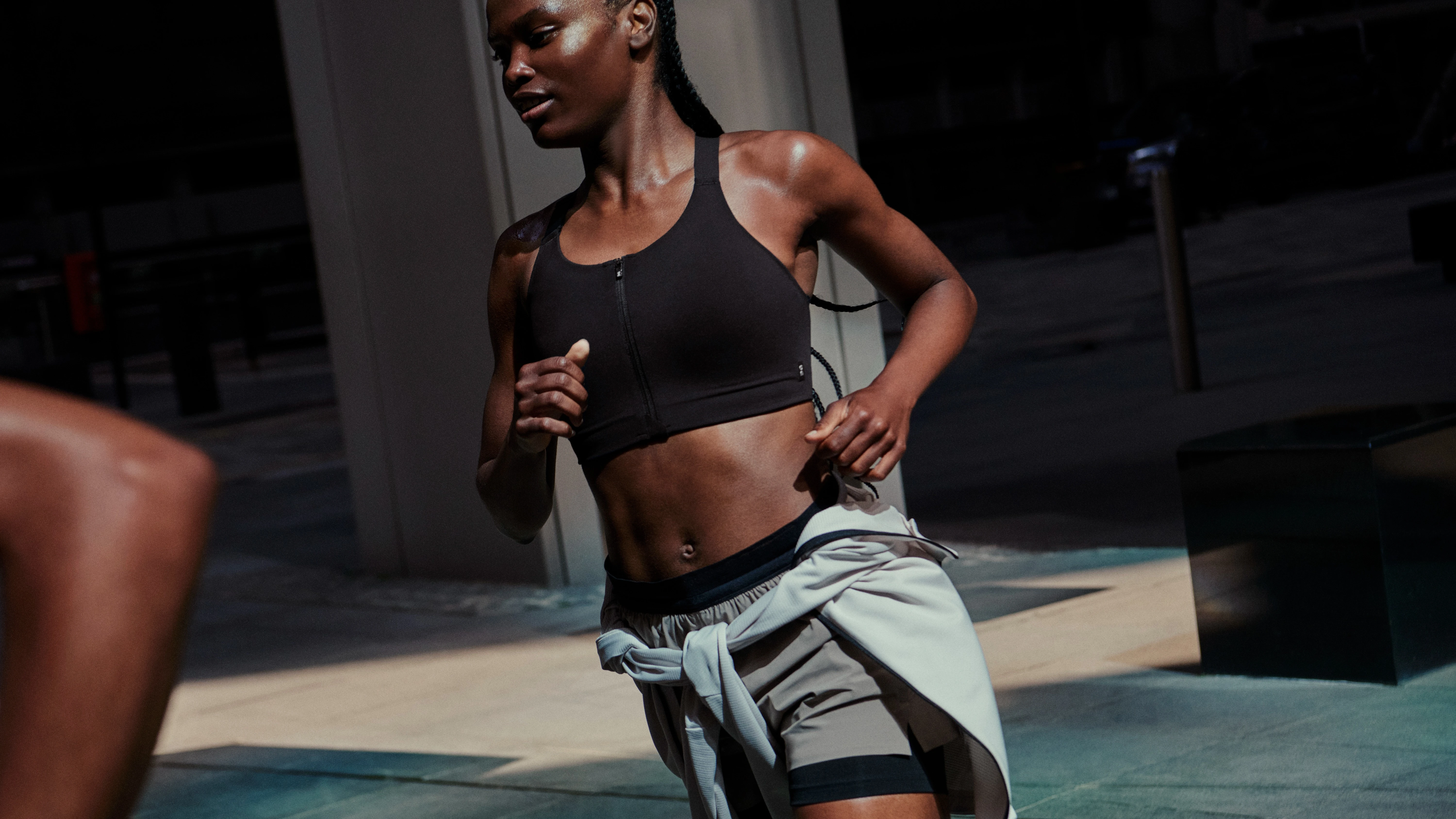 Une femme qui court en plein air, vêtue d’une brassière de sport et d’un short On, un sweat-shirt noué autour de la taille.