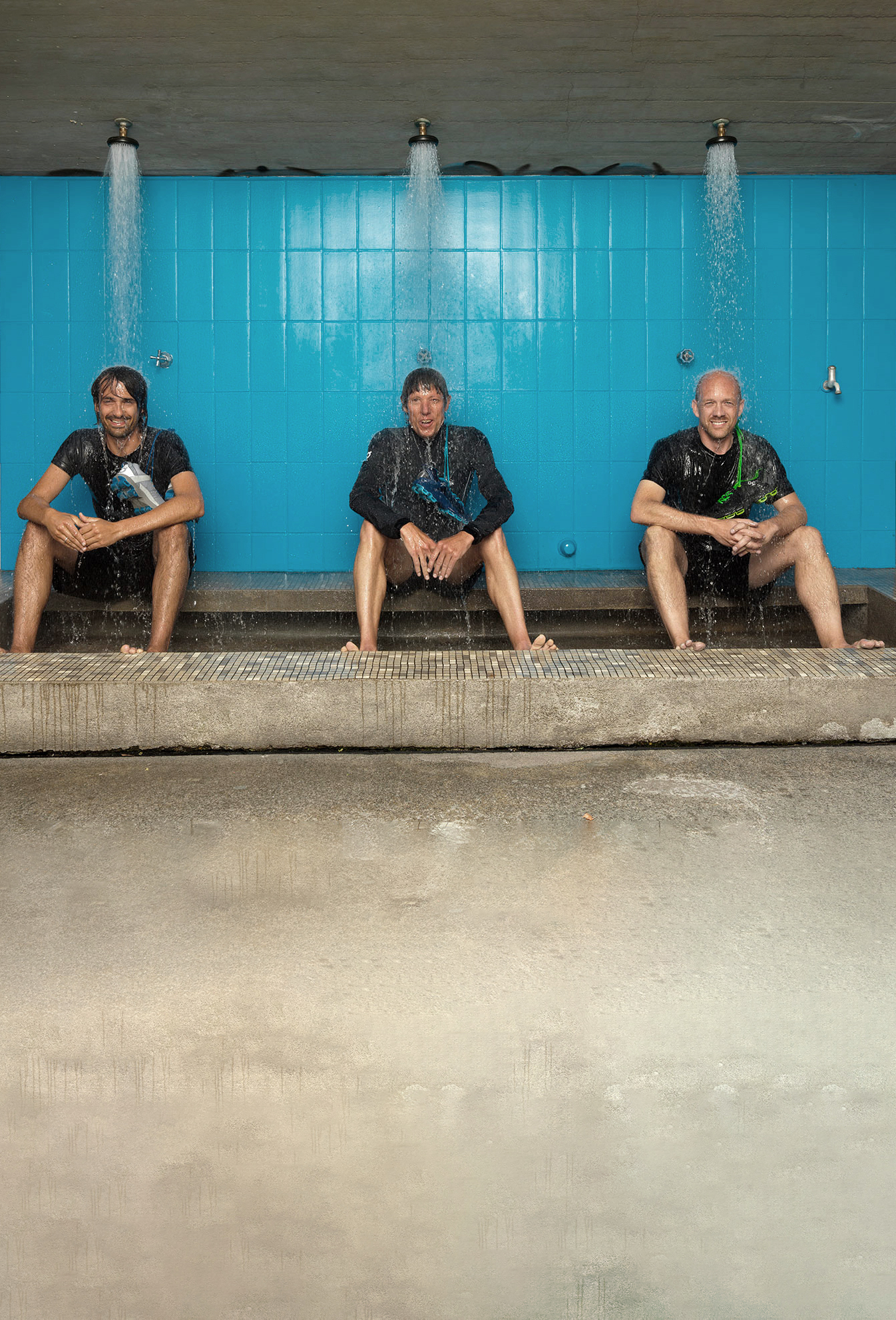 Les trois fondateurs de On sont assis dans une douche en extérieur avec de l’eau qui coule sur eux