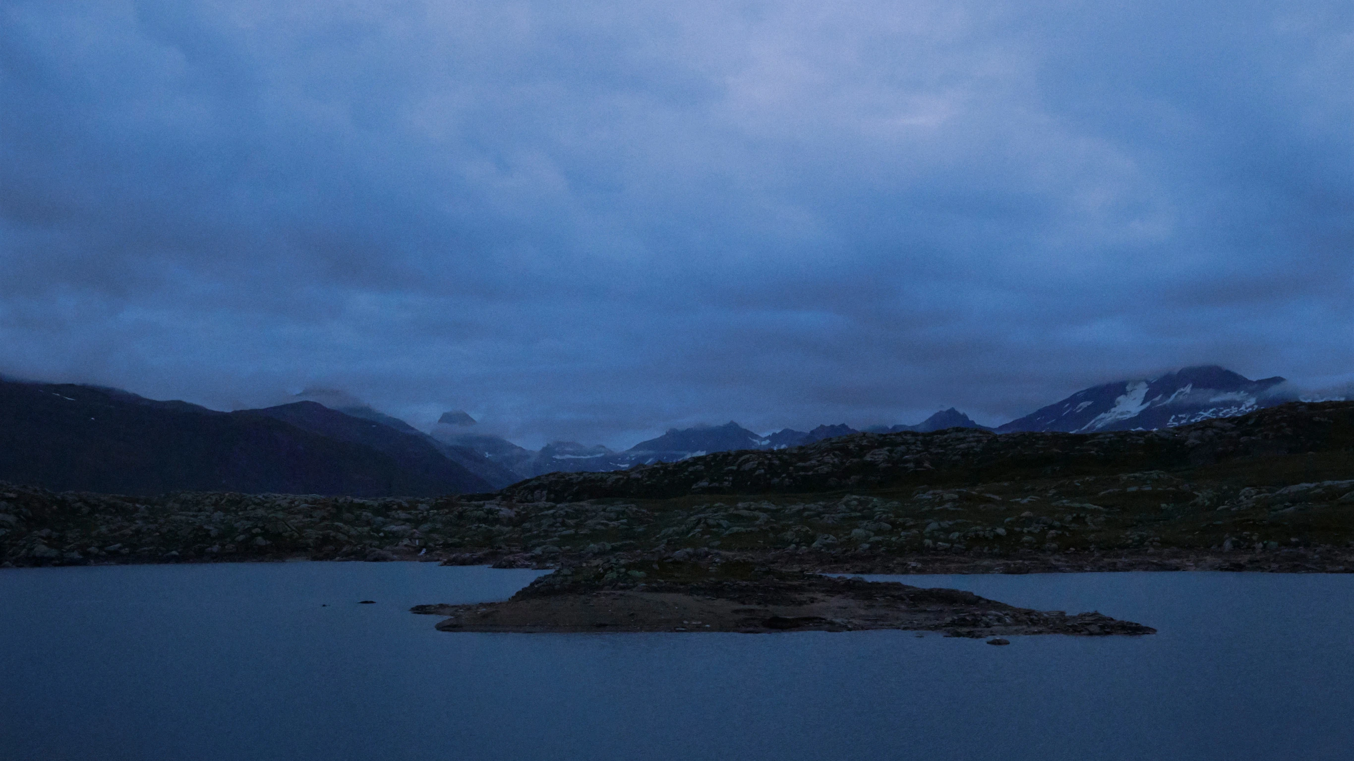 Une vue panoramique d’une journée froide, avec un lac en premier plan, des collines sombres en deuxième plan et des montagnes enneigées au loin.