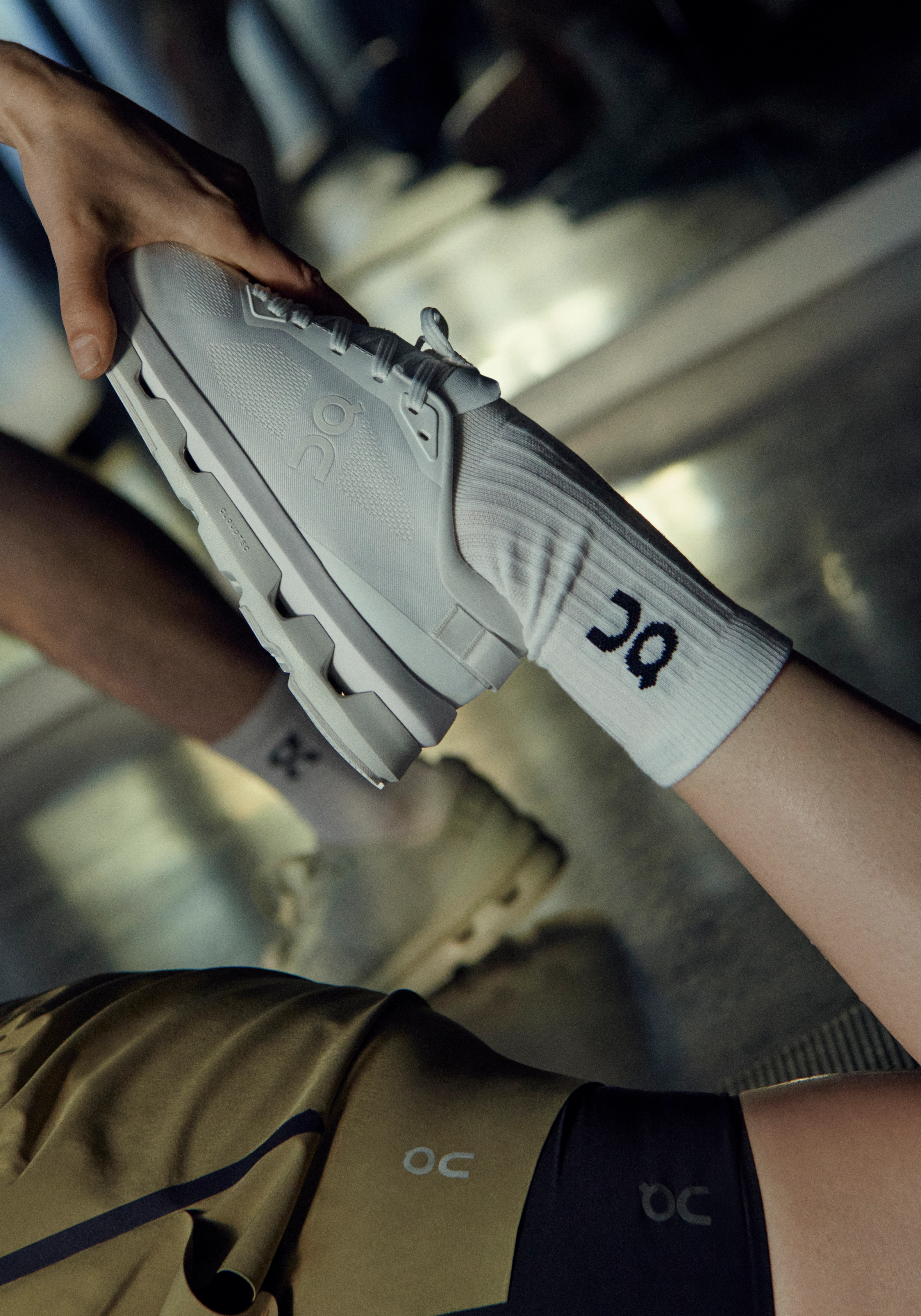 Les mains d'une personne ajustent une chaussure On grise et des chaussettes blanches. Elle porte un short vert olive, avec des équipements de sport flous en arrière-plan.