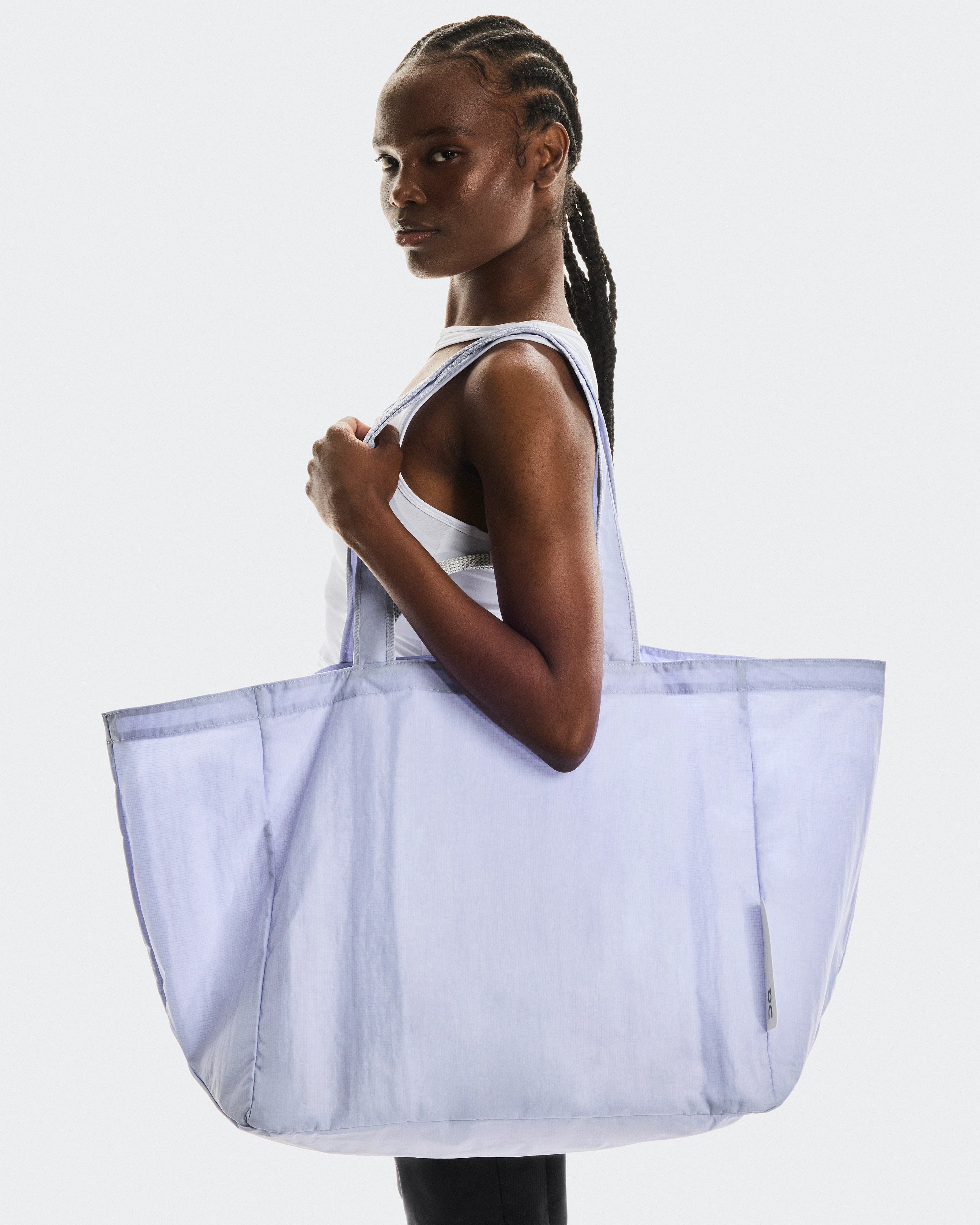 Une femme aux cheveux tressés, vêtue d’un débardeur blanc, tient un grand sac fourre-tout en mesh violet de la marque On, sur un fond gris clair.