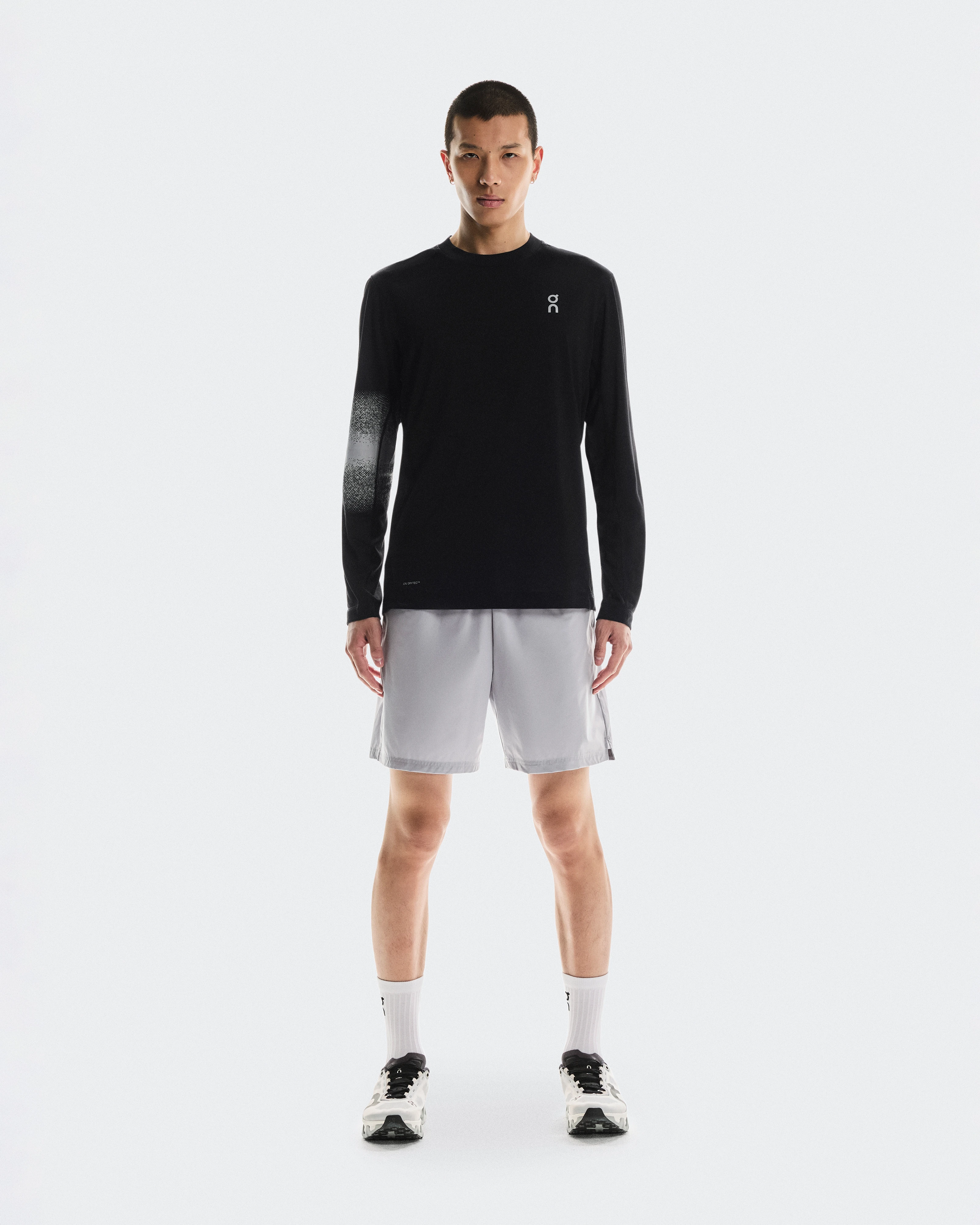 Un athlète debout, vêtu d’un t-shirt de performance à manches longues, d’un short et de chaussures de training On.