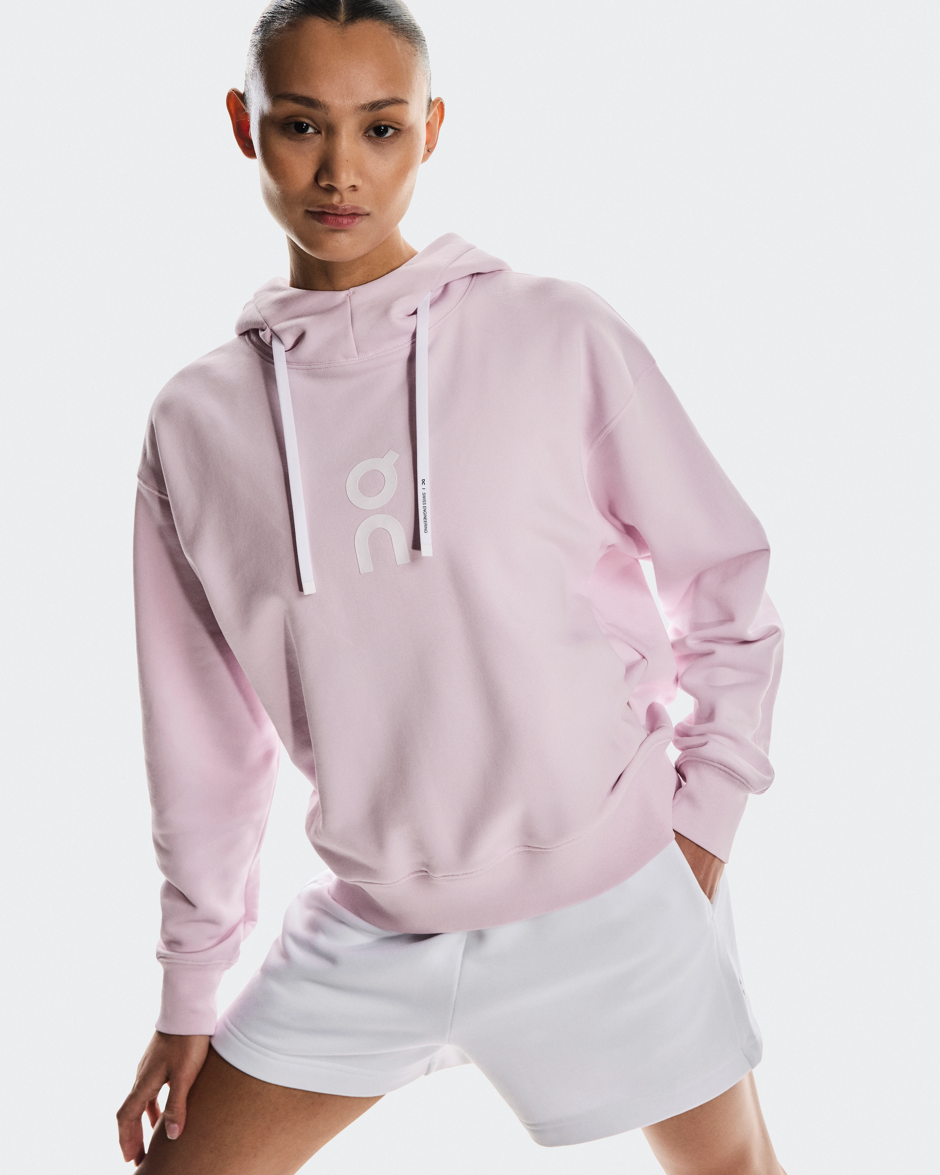 Une femme portant un sweat-shirt rose à capuche On et un short blanc, la main dans la poche.