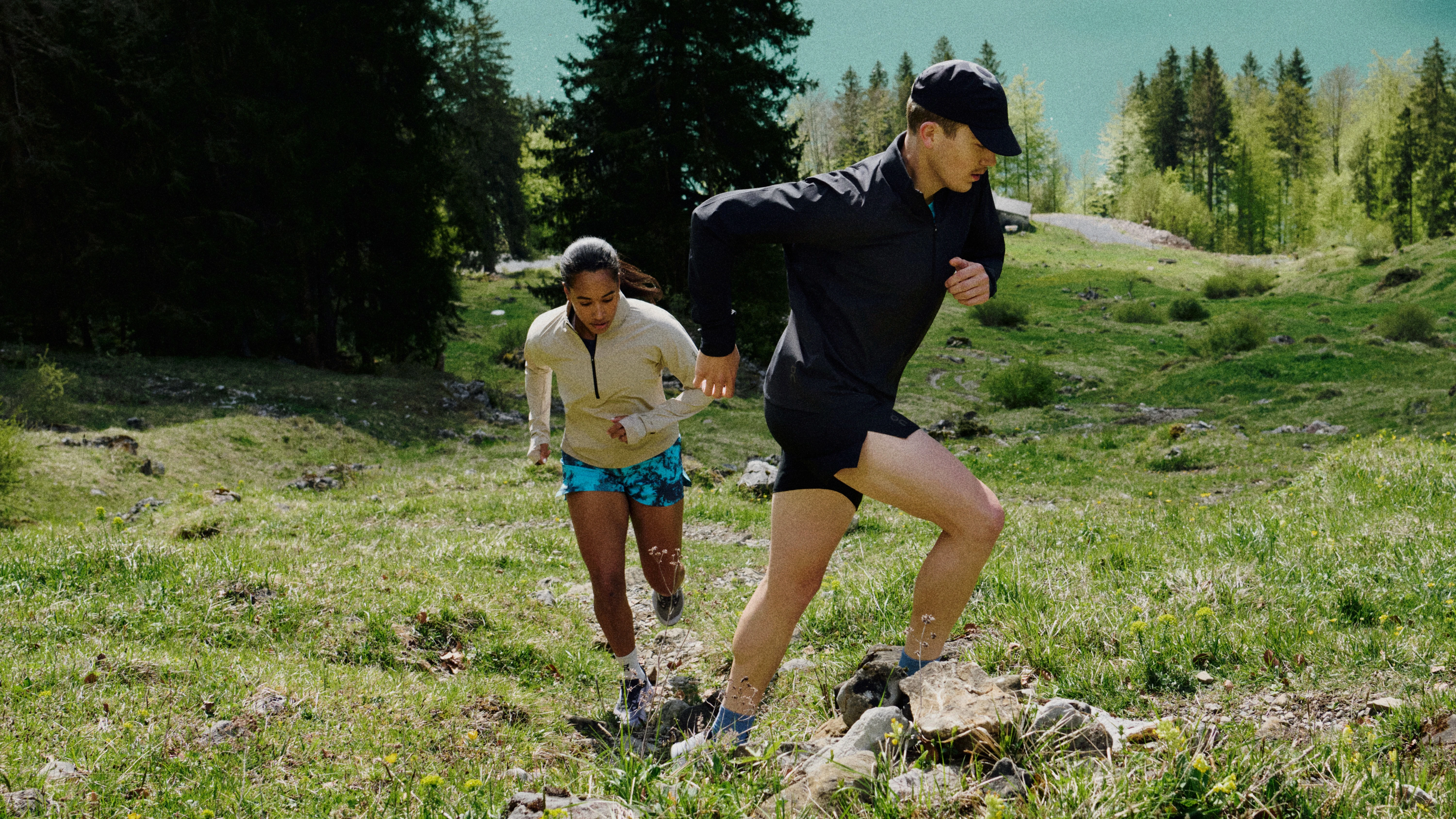 Une runneuse et un runner en tenues On courent en montée sur un sentier de montagne entourés d’herbe et d’arbres.