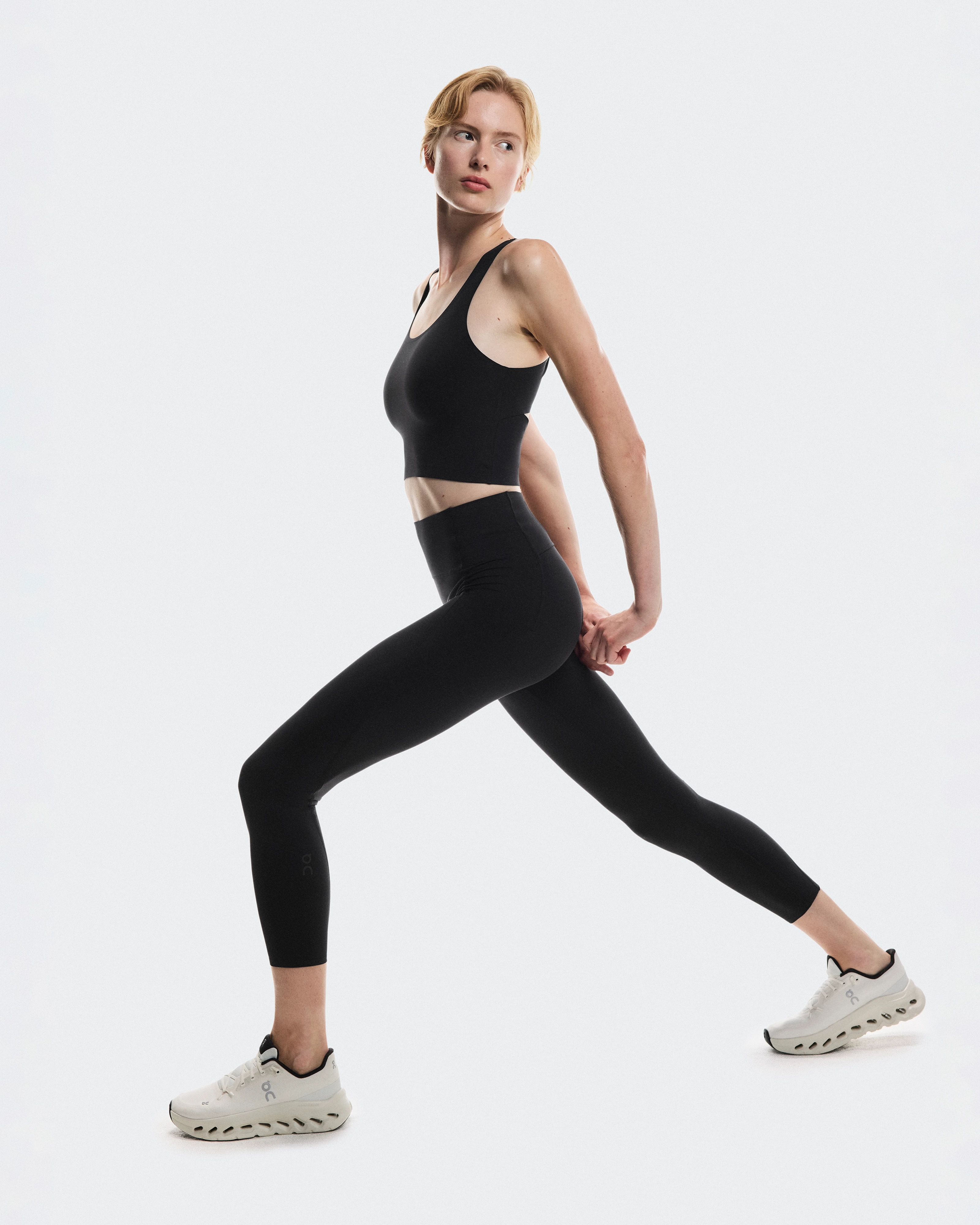 Une femme qui fait une fente, vêtue d'un débardeur court noir et d'un legging assorti de la marque On Running.