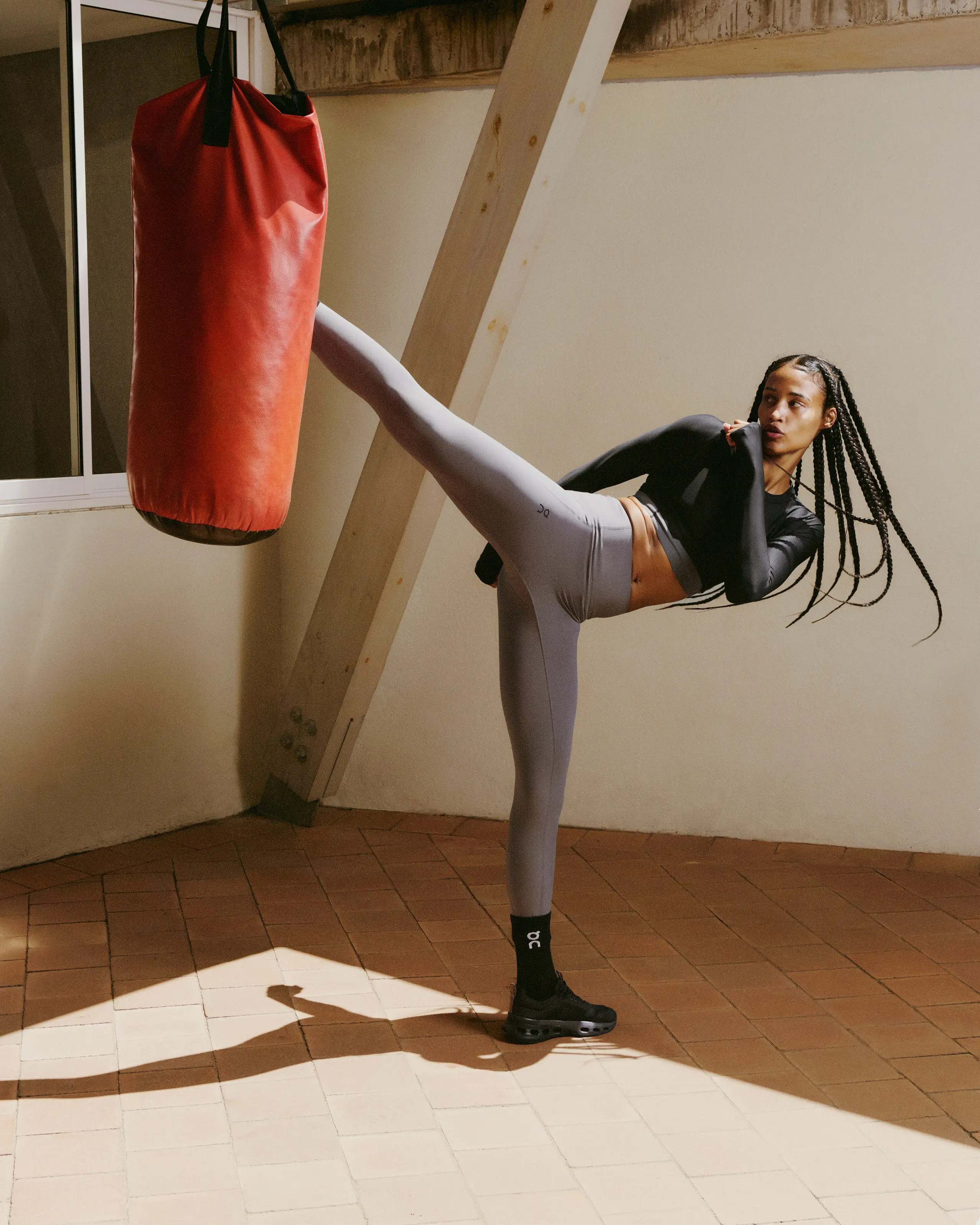 Une athlète effectue un coup de pied haut sur un sac de boxe dans une salle, vêtue de vêtements de training On.