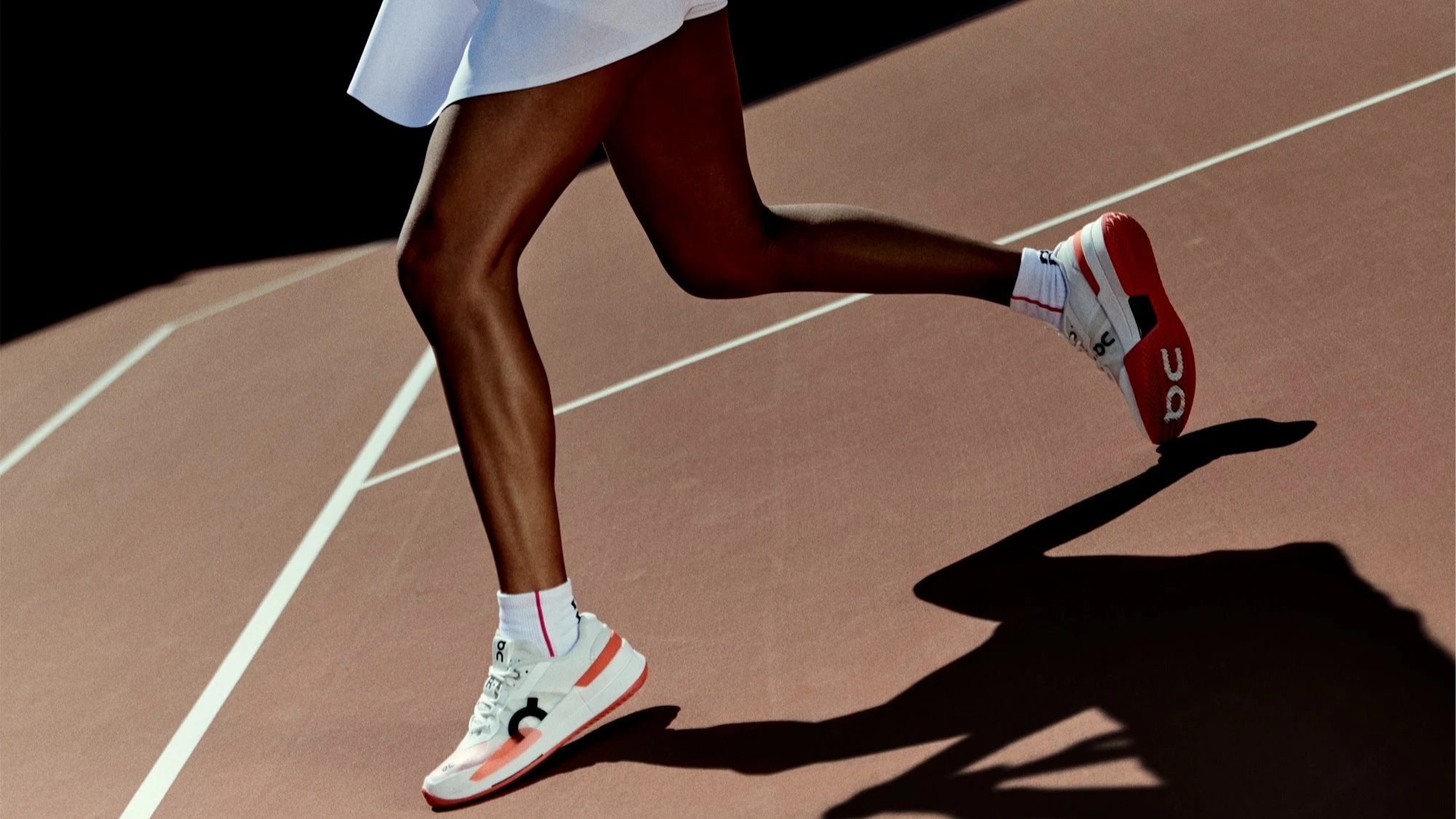 Une femme en jupe de tennis blanche court sur un court de tennis en terre battue.