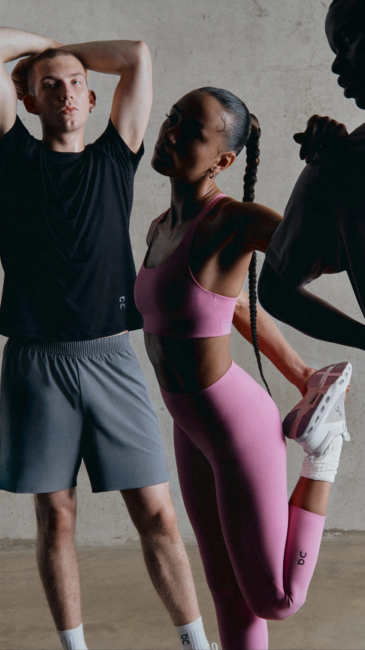 Trois athlètes en tenue de sport s’étirent avant une séance de sport en salle. Deux hommes en shorts et t-shirts ton sur ton avec une femme qui porte une brassière de sport rose et un legging.